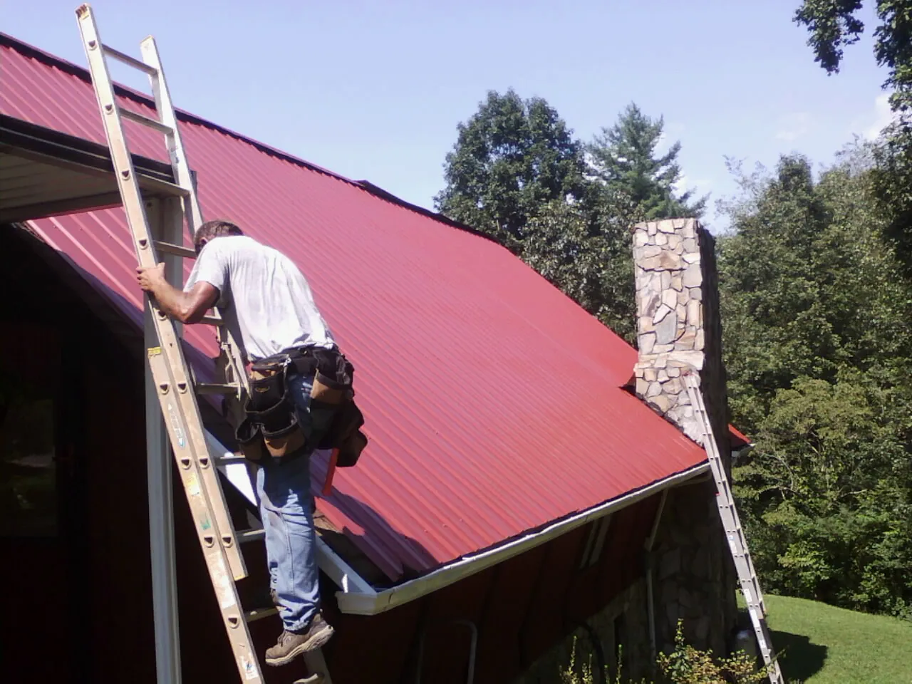 Roofer on a ladder beside a red metal roof and stone chimney — Soffit & Fascia Repair by Roofing Brothers in Centreville